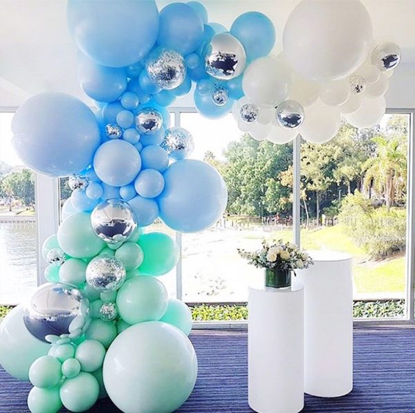 The Elegant Blue and Green Balloon Arch, offered by Valentine-Day.Shop, includes metallic silver balloons against scenic window views of trees and a lake. Two white cylindrical stands with white and yellow flower bouquets enhance the charm of this picturesque event setup.