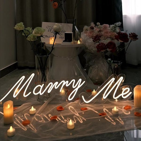 A romantic scene with the Marry Me Neon Light on sheer fabric, lit candles, and rose petals. Bouquets decorate a round table in the background, setting an ideal engagement atmosphere.