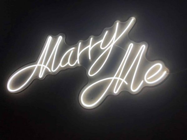 The Marry Me Script Neon Light emits a warm glow against a dark background, creating a romantic ambiance with its elegant cursive design.