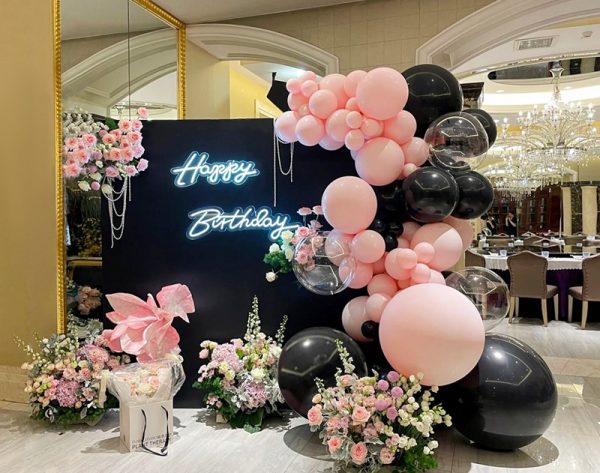 The Luxe Pink and Black Birthday Balloon Display with Floral Accents features a Happy Birthday neon sign against a dark backdrop, vibrant balloons, pink and white flowers at the base, and a transparent gift box adorned with a pink bow.