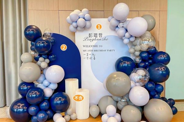 The Elegant Blue and Gray Birthday Balloon Decor Set from Valentine-day.shop features clusters of white, gray, and blue balloons with a panel saying Welcome to my 10th birthday party in English and Chinese. White cylindrical stands complete the display against a wooden wall.