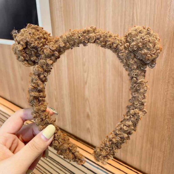 A hand holds a Teddy Bear and Sunflower Hair Band with LED, featuring fuzzy brown texture and cute bear ears, against a wooden surface.