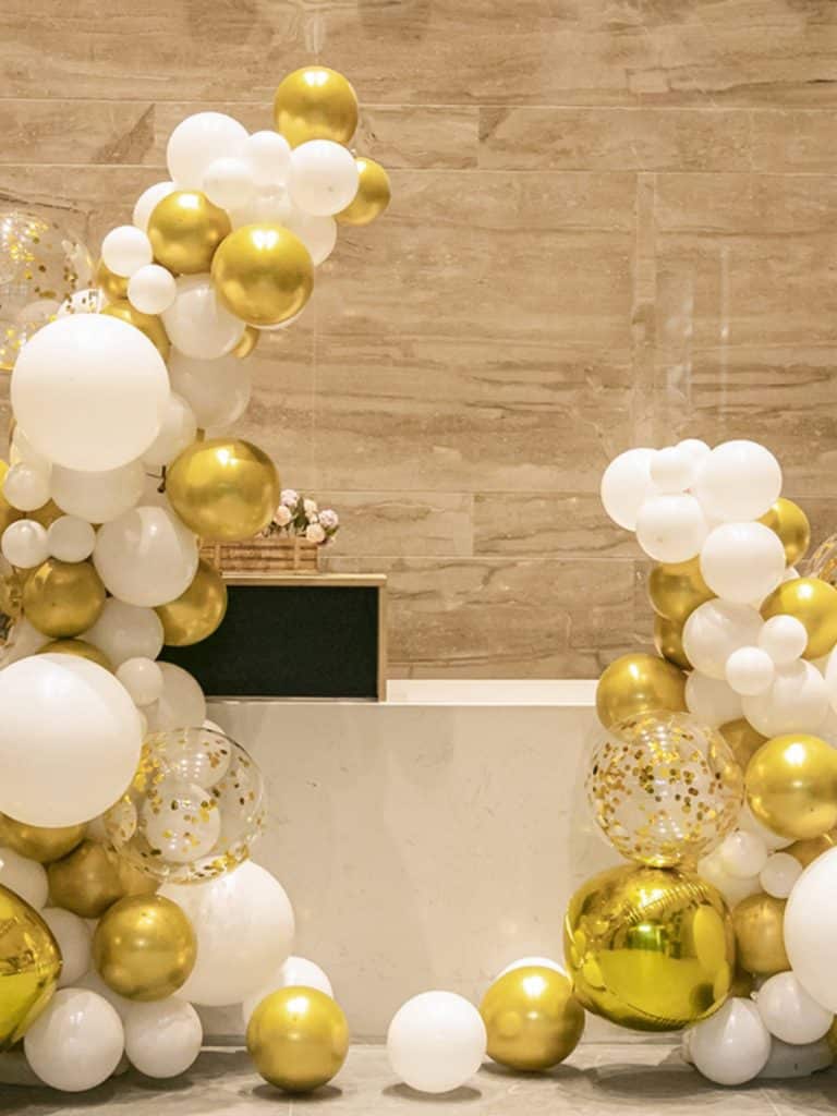The Elegant White and Gold Balloon Arch – Luxe Balloon Decorations for Events is beautifully displayed against a light beige marble wall, featuring balloons in various sizes and transparent ones filled with shimmering gold confetti.