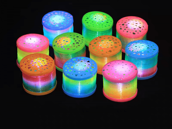 Vibrant hues of green, blue, pink, and yellow glow from colorful cylindrical lights with perforated tops on a black background. Reminiscent of a Mini Rainbow LED Bounce Spring Toy, these lights are arranged in rows for a visually striking effect.