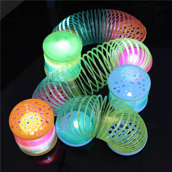 The Mini Rainbow LED Bounce Spring Toy stretches over a dark surface, its glowing lights transitioning from green to pink and blue. Its ends are capped with perforated covers to enhance the luminous display.