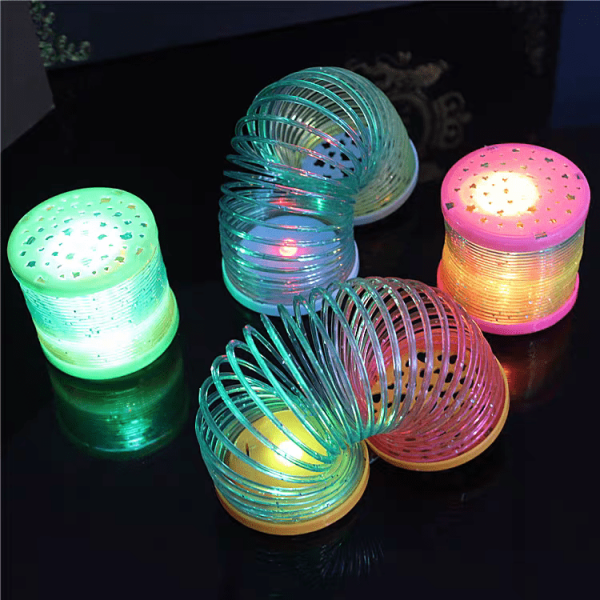 The Mini Rainbow LED Bounce Spring Toys, emanating green, blue, yellow, and pink hues, are set on a reflective surface, forming a lively display against a dark backdrop.