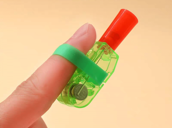 A close-up displays a hand holding an LED Cartoon Character Finger Ring, transparent green with a red tip, secured by a green strap. The button battery powers its charm against the warm background enhancing its playful allure.