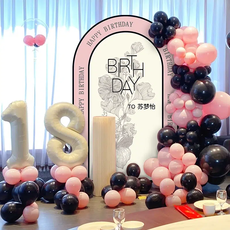 A birthday setup featuring large inflatable numbers 18 in gold, surrounded by clusters of pink and black balloons. A backdrop reads HAPPY BIRTHDAY with floral designs. The scene exudes a festive atmosphere.