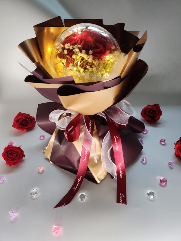 The Acrylic Ball Red Rose Bouquet with LED Lights features a clear globe containing red roses and white flowers, elegantly wrapped in shiny gold and maroon. Decorated with ribbons, it rests on a table adorned with rose petals and gems.