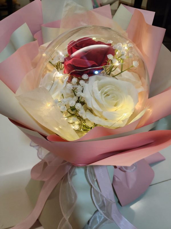 The Acrylic Ball Red and White Rose Bouquet with LED Lights features a red and white rose in a clear spherical cover, adorned with delicate white flowers, soft pink wrapping paper, warm string lights, and finished with a ribbon for an enchanting Valentines Day gift.