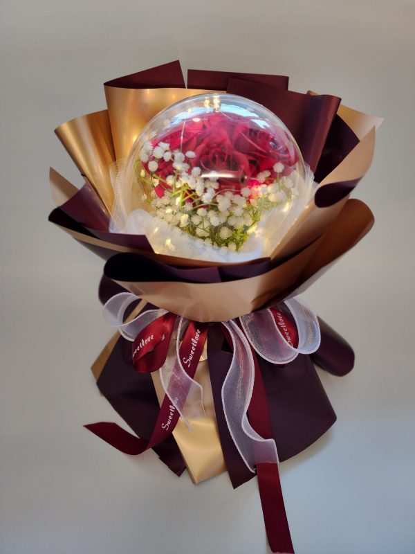 The Acrylic Ball Red Rose Bouquet with LED Lights features red roses and white flowers in a transparent sphere, wrapped in brown and gold paper, accented with a decorative bow of red and white ribbons on a neutral background.