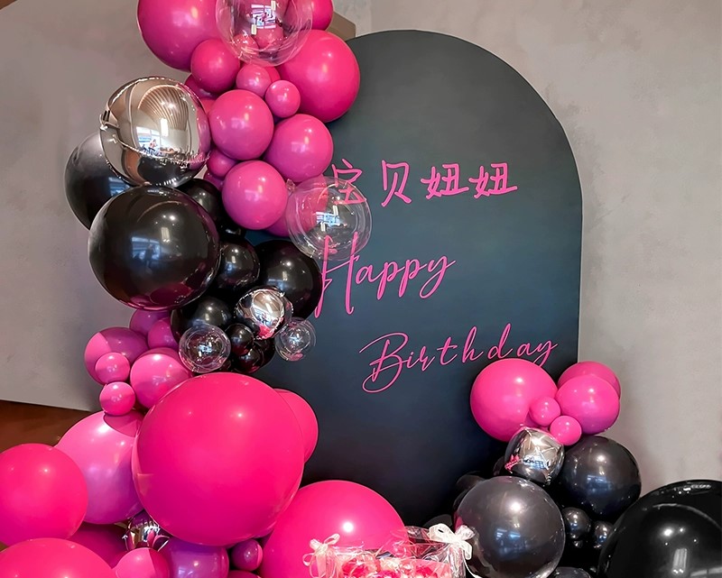 A backdrop with pink and black balloons surrounds a black panel displaying pink text that reads 宝贝妞妞 and Happy Birthday. Transparent balloons are mixed in, creating a festive birthday setting.