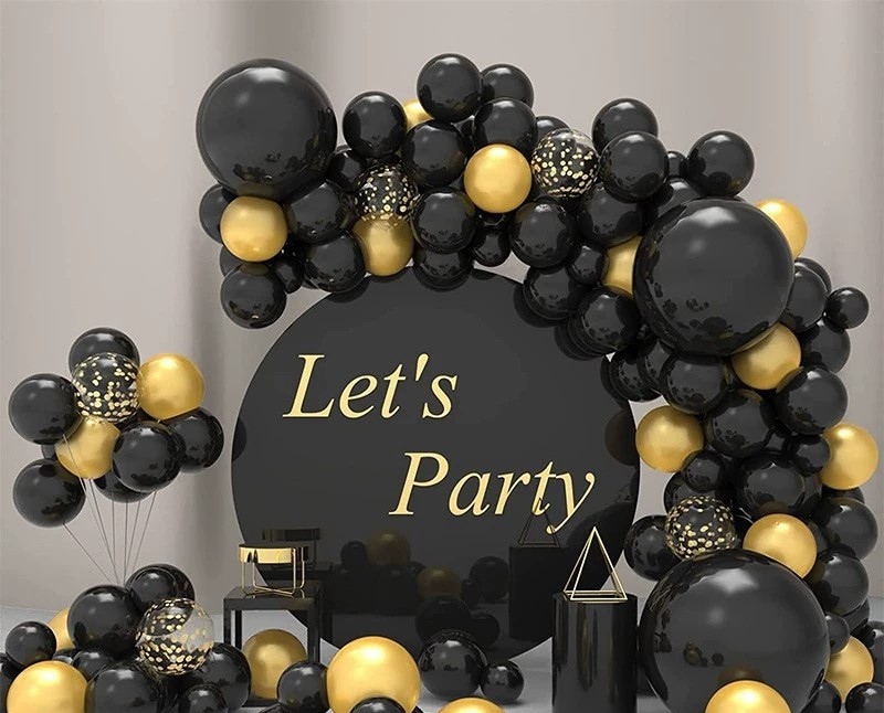 A decorative arrangement of black and gold balloons forms an arch around a black circular sign with Lets Party in gold letters. Additional balloons and geometric shapes are placed around the arch.
